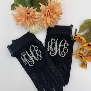 May include: Black riding gloves with a silver glitter monogram. The gloves have a textured palm and a Velcro closure. Artificial flowers in shades of peach and yellow are in the background.
