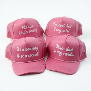 May include: Four pink trucker hats with white text. The hats have mesh backs and curved brims. The text on the hats reads: "Not slim kinda shady", "It's a bad day to be a cocktail", "I'm cool, but I cry a lot", and "Your dad is my cardio".