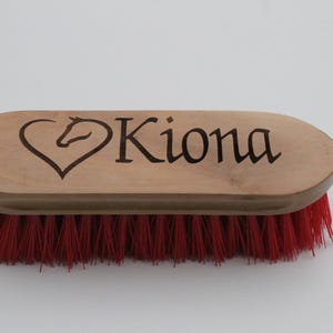 May include: Wooden horse grooming brush with red bristles. The brush features the name "Kiona" in black lettering, with a heart-shaped design incorporating a horse head. The brush is designed for equine care.