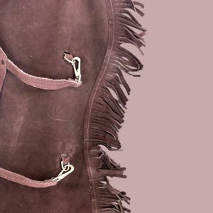 May include: Brown leather chaps with fringe detail and metal buckles. The chaps have a strap with a buckle on the front.