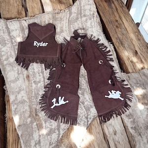 May include: Brown suede chaps with fringe and a white embroidered cowboy riding a horse on each leg. A brown suede vest with fringe and the name "Ryder" embroidered on the front.