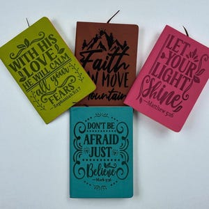 May include: Four small, colorful notebooks with inspirational quotes. The notebooks are in shades of green, brown, pink, and teal. Each notebook has a different quote in black lettering, such as "Faith Can Move Mountains."