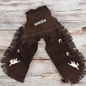 May include: Brown leather chaps with fringe and a white embroidered name "Hansen" on the vest. The chaps have a white silhouette of a rider on a horse on each leg.