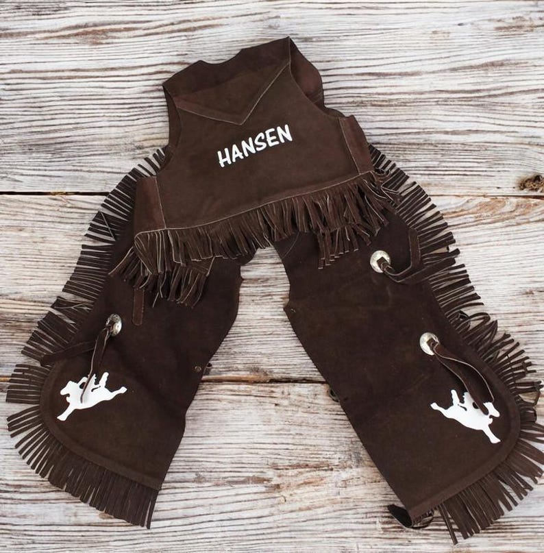 May include: Brown leather chaps with fringe and a white embroidered name "Hansen" on the vest. The chaps have a white silhouette of a rider on a horse on each leg.