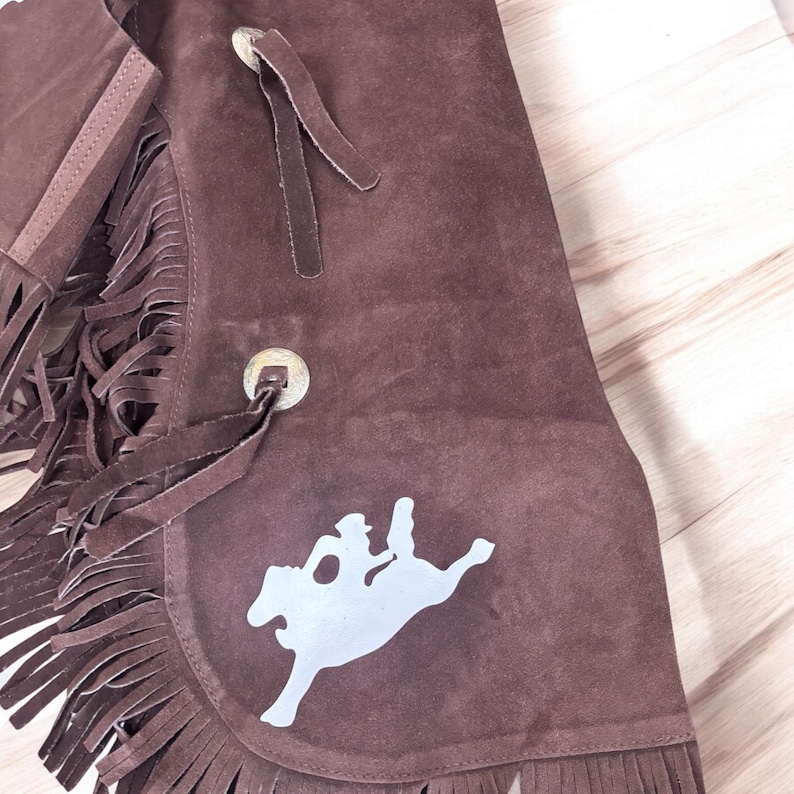 May include: Brown suede chaps with fringe detail and a silver silhouette of a rider on a horse.