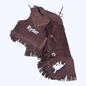 May include: Brown suede chaps and vest with fringe. The vest has the name "Ryder" embroidered on the front. The chaps have a white silhouette of a rider on a horse.