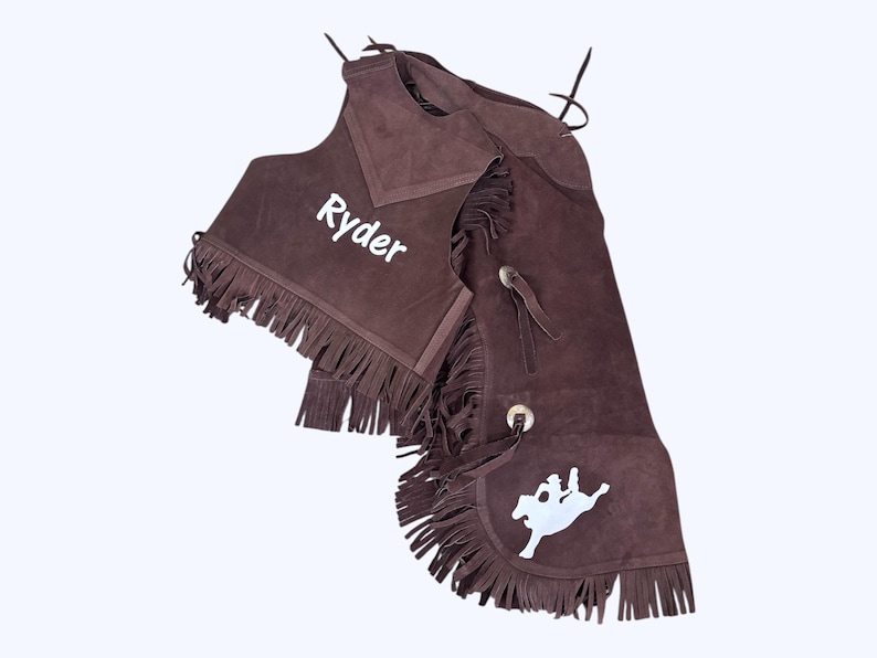 May include: Brown suede chaps and vest with fringe. The vest has the name "Ryder" embroidered on the front. The chaps have a white silhouette of a rider on a horse.