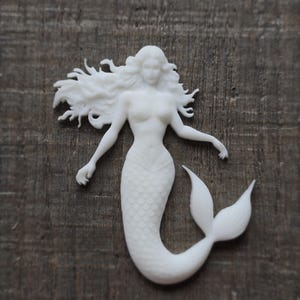 May include: A white mermaid figurine with flowing hair and a scaled tail. The mermaid has outstretched arms and is set against a dark wood grain background. The figurine is a detailed, three-dimensional sculpture.