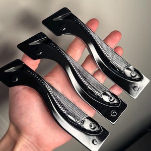 May include: Three black metal cabinet handles with a fish design. The handles have a curved shape with a textured fish pattern in the centre and are held in a hand. Each handle has a rectangular base with two screw holes.