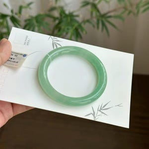 Puede incluir: Un brazalete de jade verde claro translúcido. La superficie lisa y pulida refleja la luz, mostrando las variaciones naturales de color. El brazalete se sostiene sobre una tarjeta blanca con un diseño de bambú.