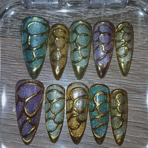 Peut inclure: Un ensemble de dix faux ongles dans un étui en plastique transparent. Les ongles sont en forme d'amande et présentent un motif chatoyant en forme d'écailles dans des tons or, sarcelle et violet, soulignés d'or. Les ongles sont disposés en deux rangées.