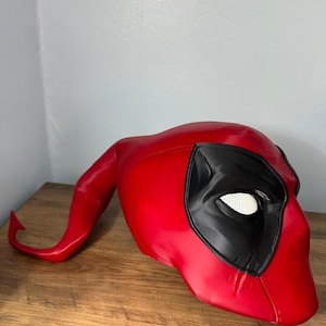 Puede incluir: Una máscara roja de Deadpool con cubiertas oculares negras y un área ocular de malla blanca. La máscara tiene una extensión curva en forma de cuerno en el lado izquierdo. La máscara está hecha de un material brillante.