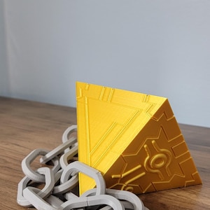May include: A golden yellow, geometric, triangular prism with a detailed design on one side, accompanied by a gray chain. The objects are displayed on a wooden surface.