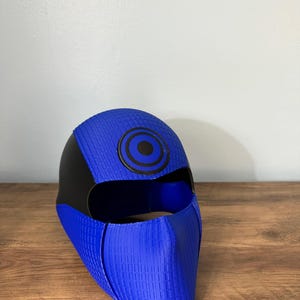 May include: A blue and black full-face mask featuring a textured grid pattern on the blue sections. The mask has a smooth black side panel and a circular black and blue target-like emblem centered on the forehead area above the eye opening.