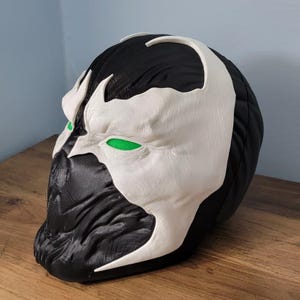 Puede incluir: Una máscara de Spawn en blanco y negro con detalles de ojos verdes. La máscara tiene una superficie texturizada y se exhibe sobre una superficie de madera. La máscara es una réplica del personaje de cómic.