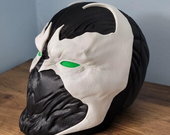 May include: A black and white Spawn mask with green eye details. The mask has a textured surface and is displayed on a wooden surface. The mask is a replica of the comic book character.