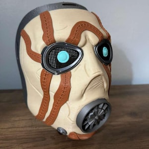 May include: A detailed mask with a cream-colored face, brown stitched accents, and black eye frames. The mask features a gray top and a gray and brown respirator. The eyes have a turquoise center.