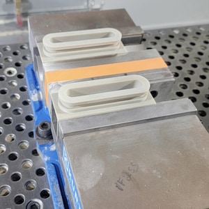 May include: Two light gray, oval-shaped, 3D-printed plastic components with a textured surface and a hollow center, secured within the metal jaws of a blue industrial machine vise. The vise features a horizontal orange strip across the metal body.