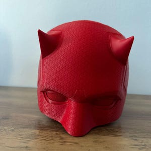 May include: A red plastic mask with horns and eye holes. The mask is designed to look like Daredevil, a superhero from Marvel Comics.