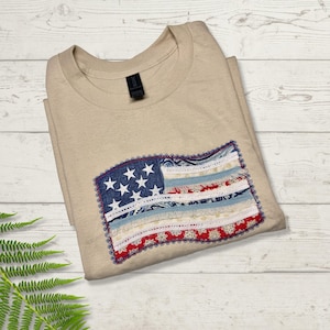 May include: Beige t-shirt featuring an American flag design. The flag is made of various fabric strips in red, white, and blue, with white stars on a blue background. The shirt is folded and lies on a light wood surface.