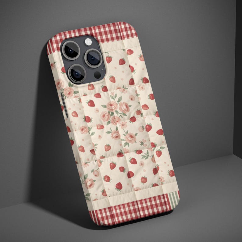 May include: A smartphone case featuring a quilted-style pattern with a cream background, red strawberries, and pink roses. The top and bottom edges have a red and white gingham border. The case has a cutout for a triple-lens camera system.
