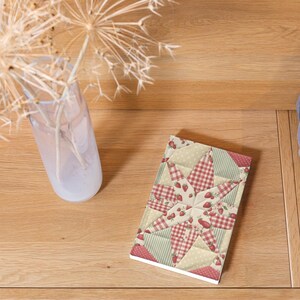 May include: A rectangular notebook featuring a quilt-inspired star pattern with red and white gingham, light green stripes, and cream polka dots, all decorated with small red strawberry illustrations across the surface of the cover.