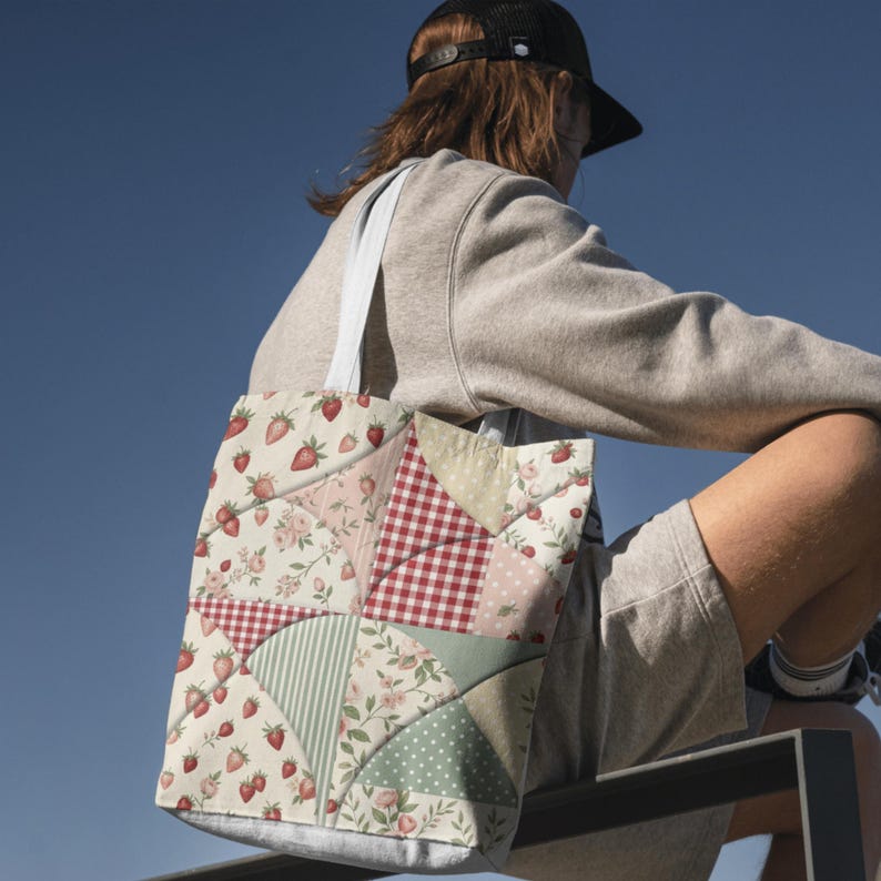 May include: A tote bag featuring a patchwork design with various patterns including red strawberries on a cream background, red and white gingham, sage green stripes, and small white polka dots on a green field, with white fabric shoulder straps.