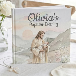 Può includere: Un libro con copertina rigida intitolato "Olivia's Baptism Blessing" con un'illustrazione ad acquerello di Gesù che tiene in braccio un bambino. Il libro è su una tovaglia bianca, accanto a un vaso di fiori bianchi e un bavaglino.