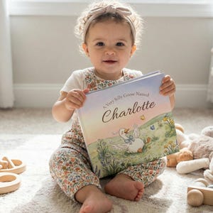 May include: A child holding a book titled "A Very Silly Goose Named Charlotte." The book cover features an illustration of a goose in a field. The child is wearing a floral print outfit and a beige headband.