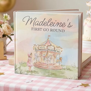 May include: A square hardcover book with a pastel watercolor illustration of a carousel, a child, and the text "Madeleine's FIRST GO ROUND" on the cover. The book is displayed on a pink and white checkered surface with decorative party items.