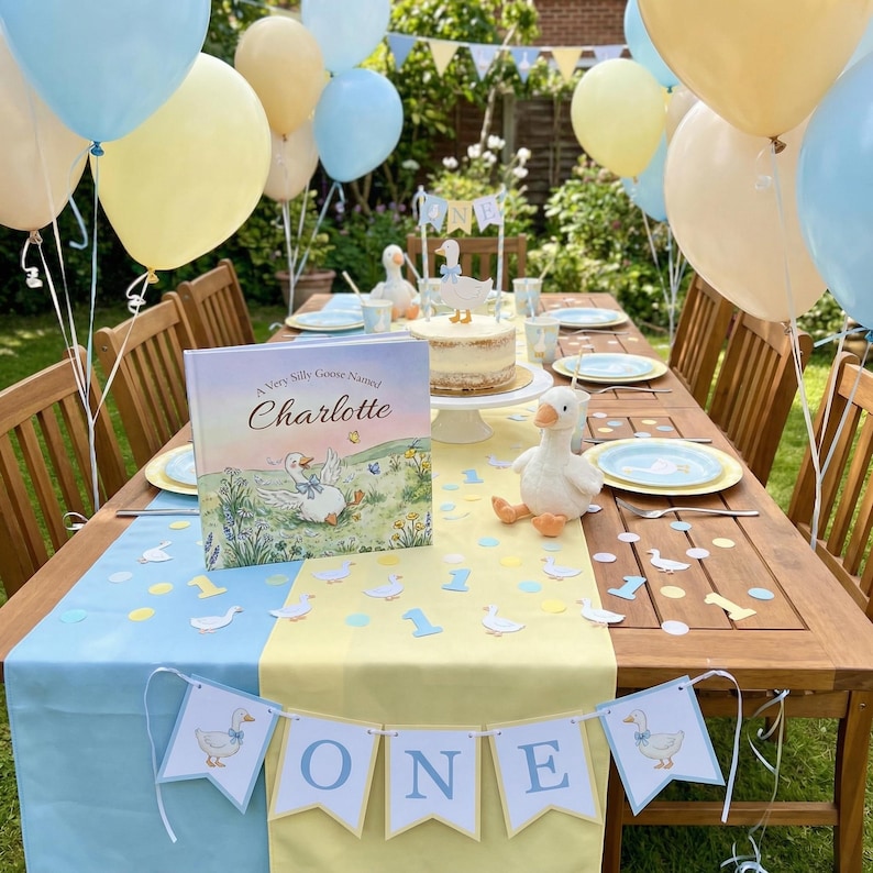 May include: A birthday party table decorated with blue, yellow, and white balloons, a cake, and a banner that says "ONE". The table is set with plates, napkins, and a book titled "A Very Silly Goose Named Charlotte".