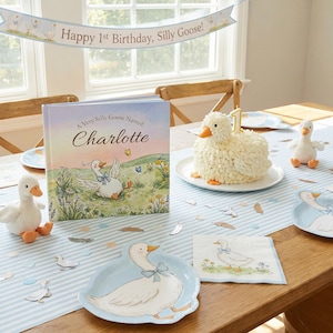 May include: A children's birthday party scene with a goose theme. Includes a cake, book, plush toys, plates, and a banner that reads "Happy 1st Birthday, Silly Goose!" The book is titled "A Very Silly Goose Named Charlotte."