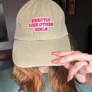 Könnte beinhalten: Eine beige Baseballkappe mit einem rosa und weißen gestickten Aufnäher mit der Aufschrift "EXACTLY LIKE OTHER GIRLS".