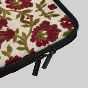 Op de afbeelding: Een crèmekleurige laptoptas met een bloemmotief in bordeauxrood en olijfgroen. Het ontwerp heeft grote, gestileerde bloemen en bladeren. De tas heeft een zwarte ritssluiting.