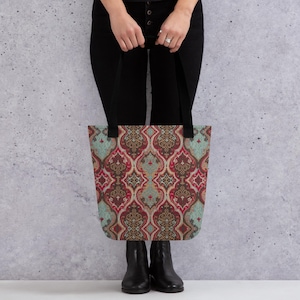 Puede incluir: Un bolso tote con un patrón colorido y ornamentado. El diseño presenta un motivo repetido en tonos rojos, marrones y turquesas. El bolso tiene asas negras y lo sostiene una persona con pantalones y botas negras.