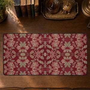 May include: A rectangular desk mat featuring a repeating floral pattern with deep red background tones and muted sage green leaves. The design displays symmetrical botanical motifs with light pink and cream-colored blossoms across the surface.