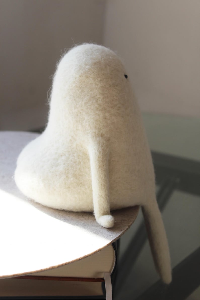 K&ouml;nnte beinhalten: Eine wei&szlig;e Filzskulptur einer Kreatur mit einem runden K&ouml;rper und langen Armen. Die Kreatur sitzt auf einer braunen Oberfl&auml;che.