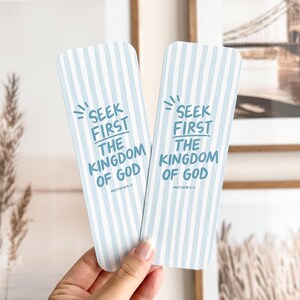 May include: Two light blue and white striped bookmarks with the text "Seek First The Kingdom of God" in blue. The bookmarks are held in a hand, with framed pictures in the background.