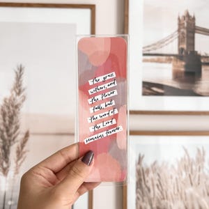 May include: A pink and gray watercolor bookmark with the quote "The grass withers, and the flower falls, but the word of the Lord remains forever."