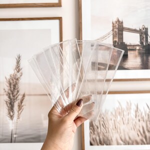 May include: A set of clear plastic bookmark sleeves, held in a hand against a background of framed pictures.