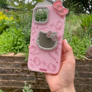 Pu&ograve; includere: Custodia per telefono rosa con motivi di Hello Kitty e un fiocco decorativo. La custodia ha un bordo trasparente con strass attorno alla fotocamera e allo specchio. Il testo "HELLO KITTY" e "AUTUMN" sono visibili.