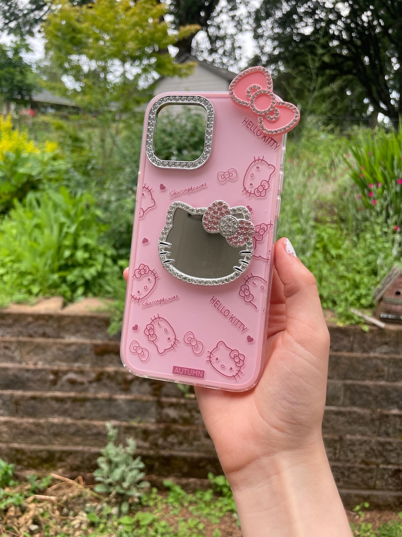 Pu&ograve; includere: Custodia per telefono rosa con motivi di Hello Kitty e un fiocco decorativo. La custodia ha un bordo trasparente con strass attorno alla fotocamera e allo specchio. Il testo "HELLO KITTY" e "AUTUMN" sono visibili.