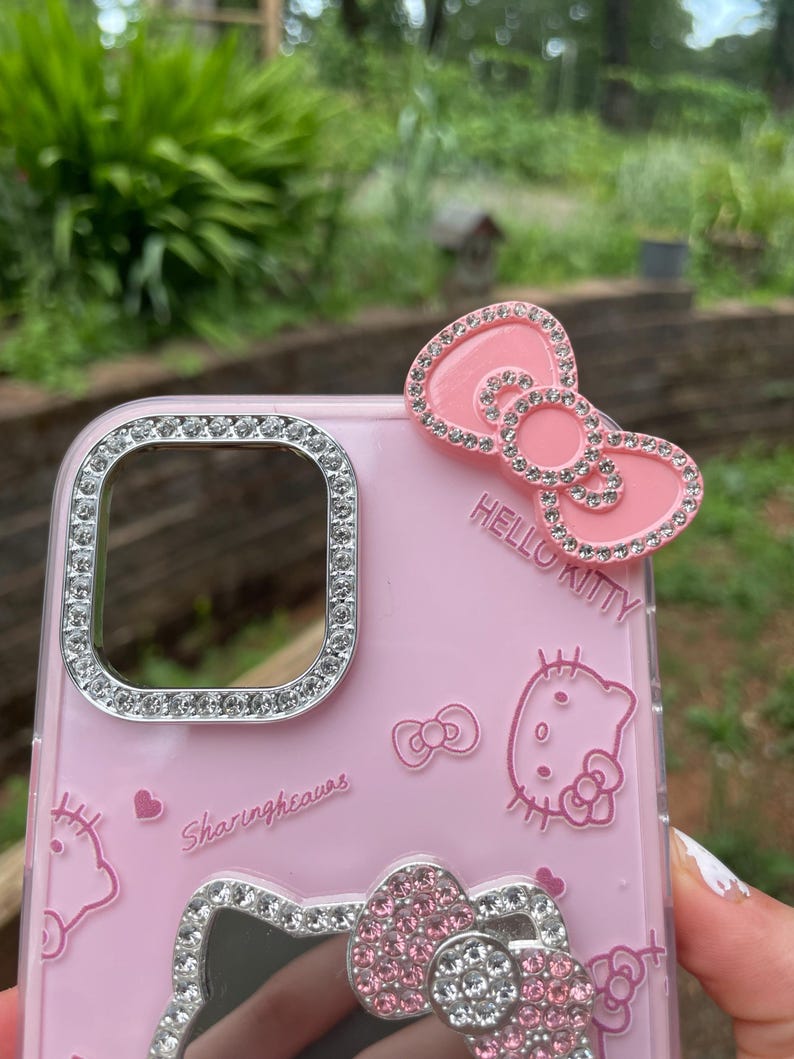 Pu&ograve; includere: Custodia per telefono rosa con un design di Hello Kitty. La custodia presenta un fiocco con strass e le parole "Hello Kitty". L'obiettivo della fotocamera e il fiocco inferiore sono anch'essi decorati con strass. La custodia ha le parole "Sharingheaves".