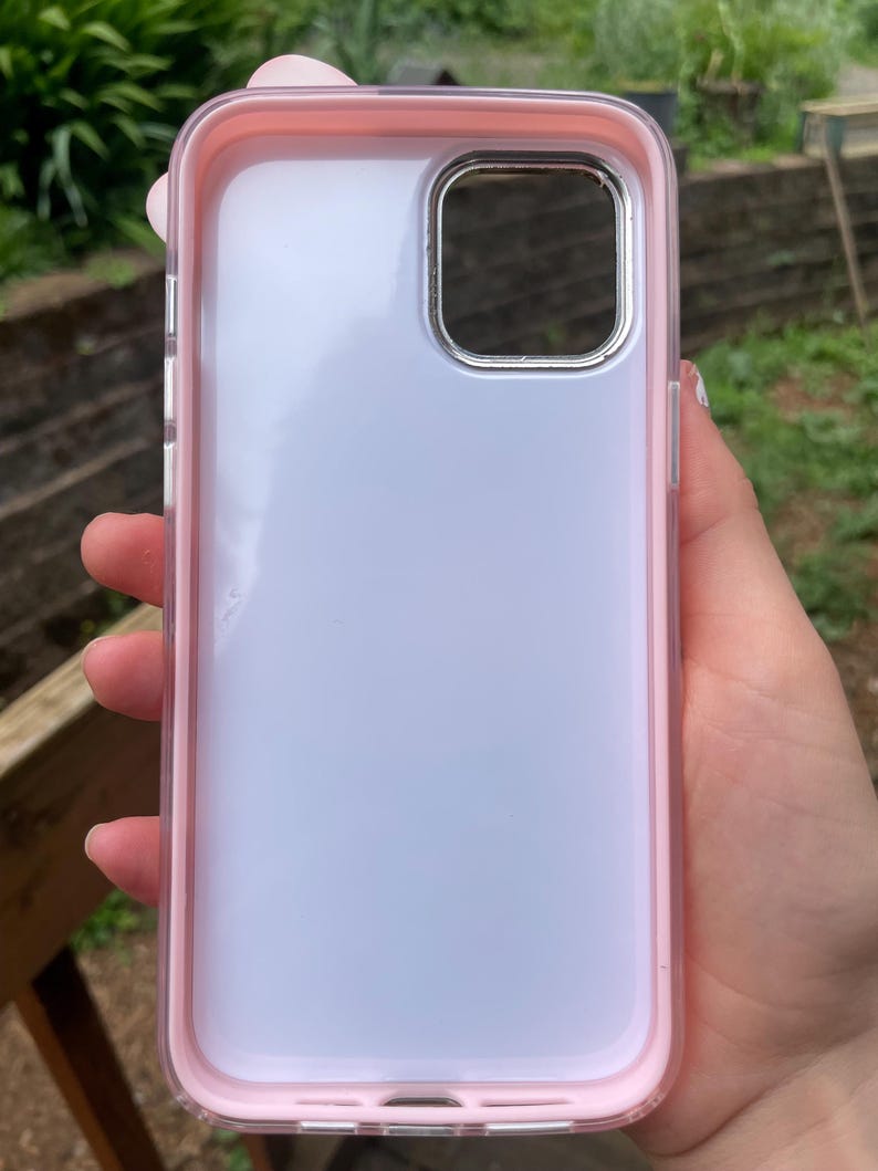 Pu&ograve; includere: Una custodia trasparente per telefono con bordo rosa. La custodia ha un ritaglio argentato per la fotocamera e un retro trasparente. La custodia del telefono &egrave; tenuta in mano da una persona.
