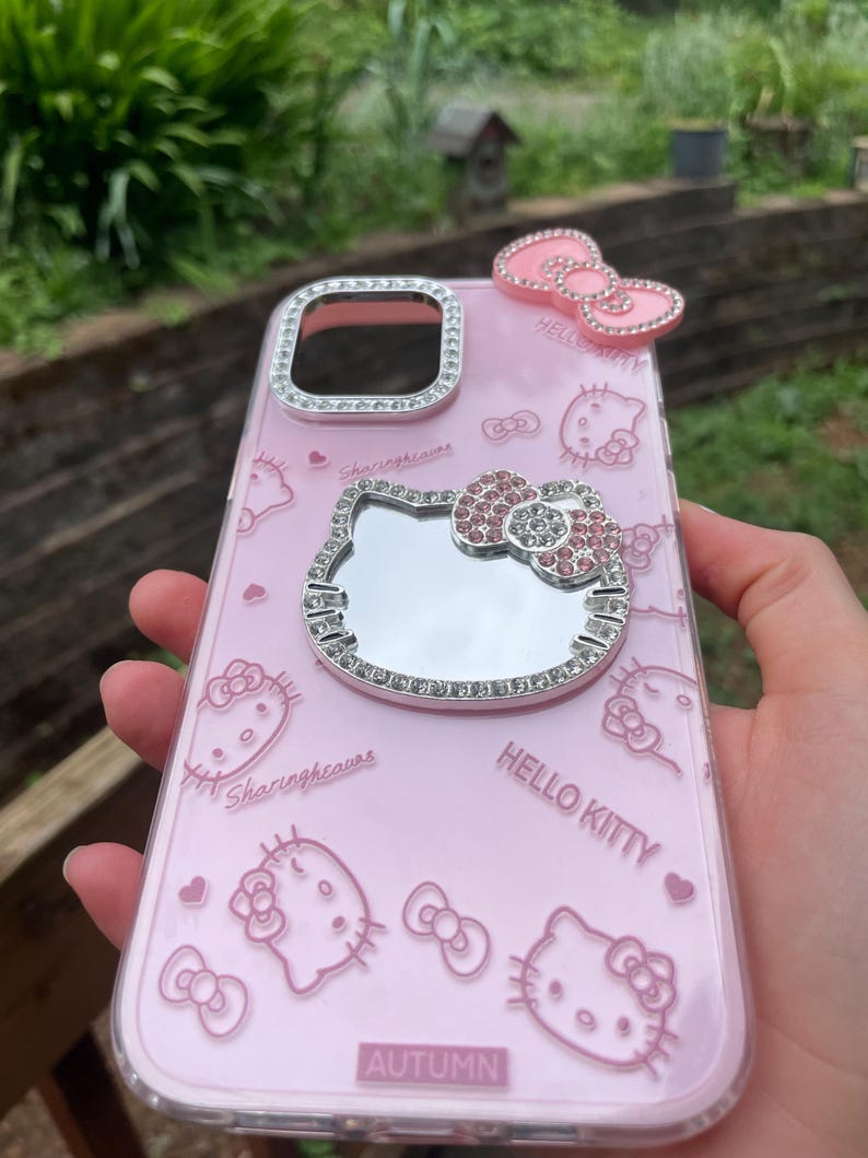 Pu&ograve; includere: Custodia per telefono Hello Kitty rosa con specchio e fiocchi. La custodia ha un bordo trasparente con finiture in strass argentati attorno alla fotocamera e allo specchio. La custodia ha le parole "Hello Kitty" e "Autumn".