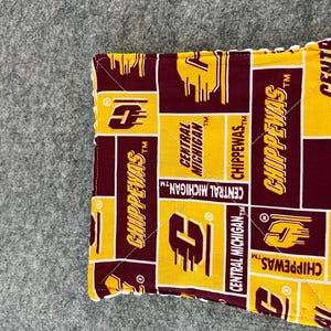 May include: A square, quilted fabric item featuring a repeating pattern of maroon and gold rectangles. The rectangles display the text "CHIPPEWAS" and "CENTRAL MICHIGAN" in a bold, collegiate font. The item is likely a coaster or a similar decorative piece.