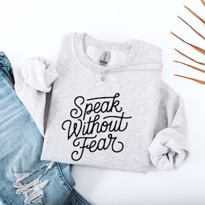 May include: A light gray sweatshirt with the words "Speak Without Fear" in black script. The sweatshirt is folded, with the sleeves rolled up. A silver necklace with a small pendant is draped across the front. Blue jeans are visible in the background.