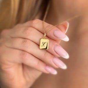 Può includere: Collana con ciondolo in oro e un ciondolo quadrato con la lettera 'K' in scrittura corsiva. Il ciondolo ha un bordo testurizzato.