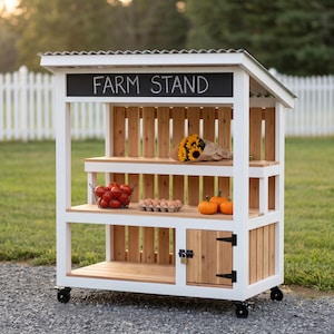 May include: A white and natural wood farm stand on wheels with a corrugated metal roof. The structure features a black sign with the white text FARM STAND, two open shelves holding a wire basket of tomatoes, eggs, and pumpkins, and a small cabinet.