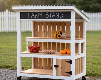 May include: A white and natural wood farm stand on wheels with a corrugated metal roof. The structure features a black sign with the white text FARM STAND, two open shelves holding a wire basket of tomatoes, eggs, and pumpkins, and a small cabinet.
